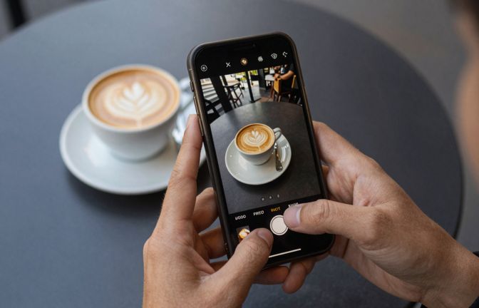 Overhead view of hands holding a phone above a cafe table to photograph a flat white and a pastry